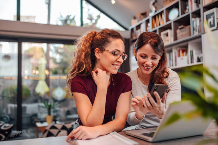 Mujeres sonriendo viendo un celular y con un portátil enfrente